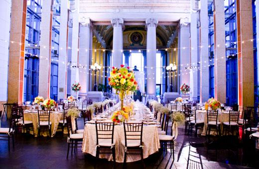 A grand banquet hall is elegantly decorated for a formal event. Long tables with white tablecloths are set with glasses and cutlery. Colorful floral centerpieces and string lights create a festive ambiance under towering columns.