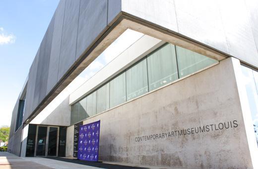 A modern concrete building with large windows, displaying the text "Contemporary Art Museum St. Louis" on its facade. A blue banner with white text is placed near the entrance.