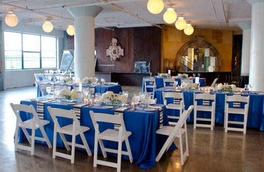 A spacious event room with large windows features tables covered in blue and white tablecloths, set for a banquet. White chairs surround the tables, which are decorated with floral centerpieces and candles. Overhead, round lights illuminate the space.