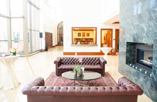 A spacious lounge with leather sofas arranged around a glass coffee table on a patterned rug. A marble fireplace is on the right, and a bar is visible in the background. Large windows provide natural light.