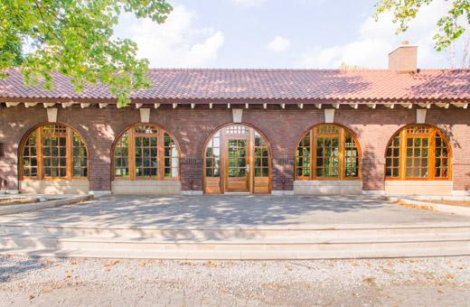 A long, brown brick building with a red-tiled roof and multiple arched wooden doorways. The windows and doors have glass panes. Sunlight filters through surrounding green trees, casting shadows on a stone walkway.