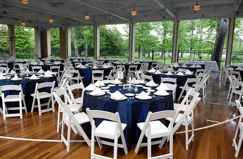 A dining area with round tables covered in dark blue tablecloths, each set with white napkins and plates. White folding chairs surround the tables. The venue has wooden floors and large windows with a view of green trees outside.