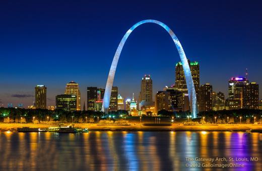 Night view of the St. Louis skyline with the Gateway Arch illuminated. The reflection of the city lights shimmers on the water in the foreground. Buildings are lit up, creating a vibrant urban scene against a dark blue sky.