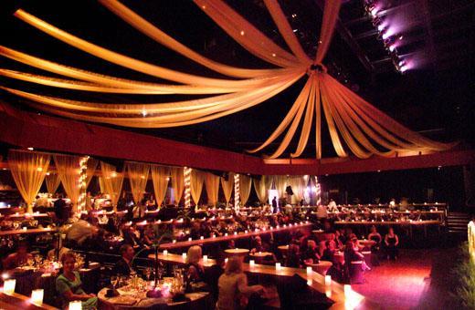 A dimly lit banquet hall with elegant drapery hanging from the ceiling. The room is filled with guests seated at long tables, enjoying a formal dinner under warm, ambient lighting. Candles are placed on the tables, enhancing the atmosphere.