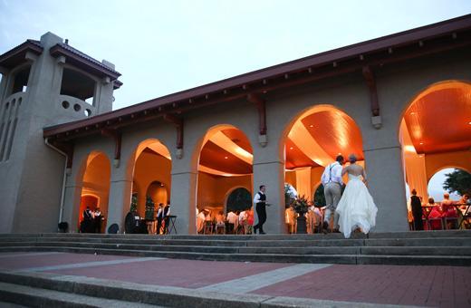 A well-lit event venue with arched doorways, featuring a bride and groom entering. Guests are mingling inside the warmly illuminated space. The building has a distinct architectural design with a prominent tower on the left.