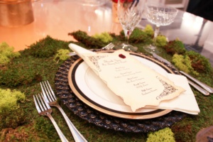 A table setting with an ornate black and white plate, silver cutlery, crystal glassware, and a decorative menu with a red wax seal. The table is adorned with a green moss centerpiece, creating an elegant and natural look.
