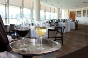 A waiter holds a tray with a glass of red wine and a glass of white wine in an elegant, sunlit dining room with white tablecloths and neatly arranged chairs. The room features large windows and framed artwork on the walls.