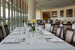 A well-lit dining room with tables set for an event. Each table is covered with a white tablecloth and adorned with yellow flowers in small vases. Wine glasses and cutlery are neatly arranged. Large windows line one side of the room, allowing natural light in.