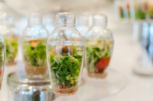 A row of clear plastic containers resembling cocktail shakers, filled with colorful layered salads, including greens and vegetables. They are displayed on a transparent tiered stand against a soft-focus background.