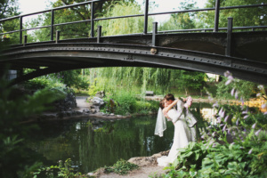 A couple in wedding attire shares a kiss beneath a black metal bridge. They stand by the edge of a lush, green pond with trees and plants surrounding them, reflecting a serene and romantic atmosphere.