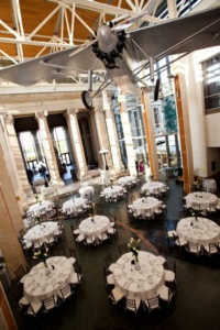 A grand hall set up for an event with round tables covered in white tablecloths and chairs arranged around them. An airplane is suspended from the ceiling. The space has large columns and windows, creating a bright, open atmosphere.