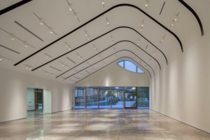 A modern, empty gallery with a high, curved ceiling features recessed lighting. The polished concrete floor reflects the natural light from large glass windows displaying an outdoor scene with trees and a staircase. A triangular window adds architectural interest.