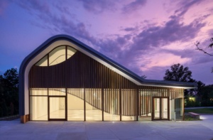 Modern building with curved roof design, featuring large windows and vertical wooden slats. The structure is illuminated from within, set against a vibrant sunset sky with purple and pink hues. Trees are visible in the background.