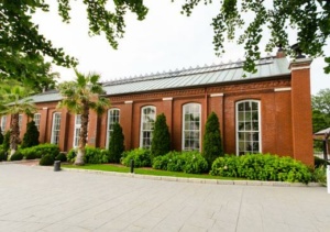 A historic brick building with tall arched windows is surrounded by lush greenery and palm trees. The foreground features a paved walkway under a clear sky.