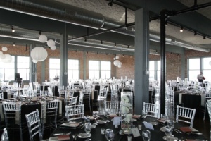 A loft-style banquet hall set up for an event, featuring round tables with black tablecloths, white flower centerpieces, and clear chairs. Large windows and hanging paper lanterns create a bright and elegant atmosphere.