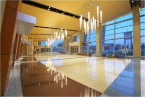 Spacious, modern lobby with high wooden ceilings, large windows, and polished floors. Numerous hanging pendant lights illuminate the space. A few chairs are arranged near the windows, with views of an outside structure visible.