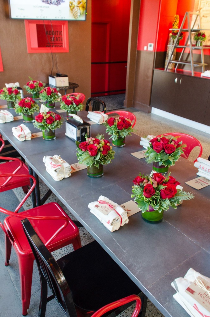 A long table set for a gathering, featuring small vases of red roses and greenery as centerpieces. Each place setting includes a menu and a neatly folded napkin. Red and black chairs surround the table in a warmly lit restaurant interior.