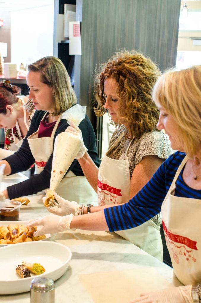 Three women wearing aprons are decorating pastries at a table. They are using piping bags to add cream. A plate with pastries is in the foreground. The setting appears to be a kitchen or cooking class.