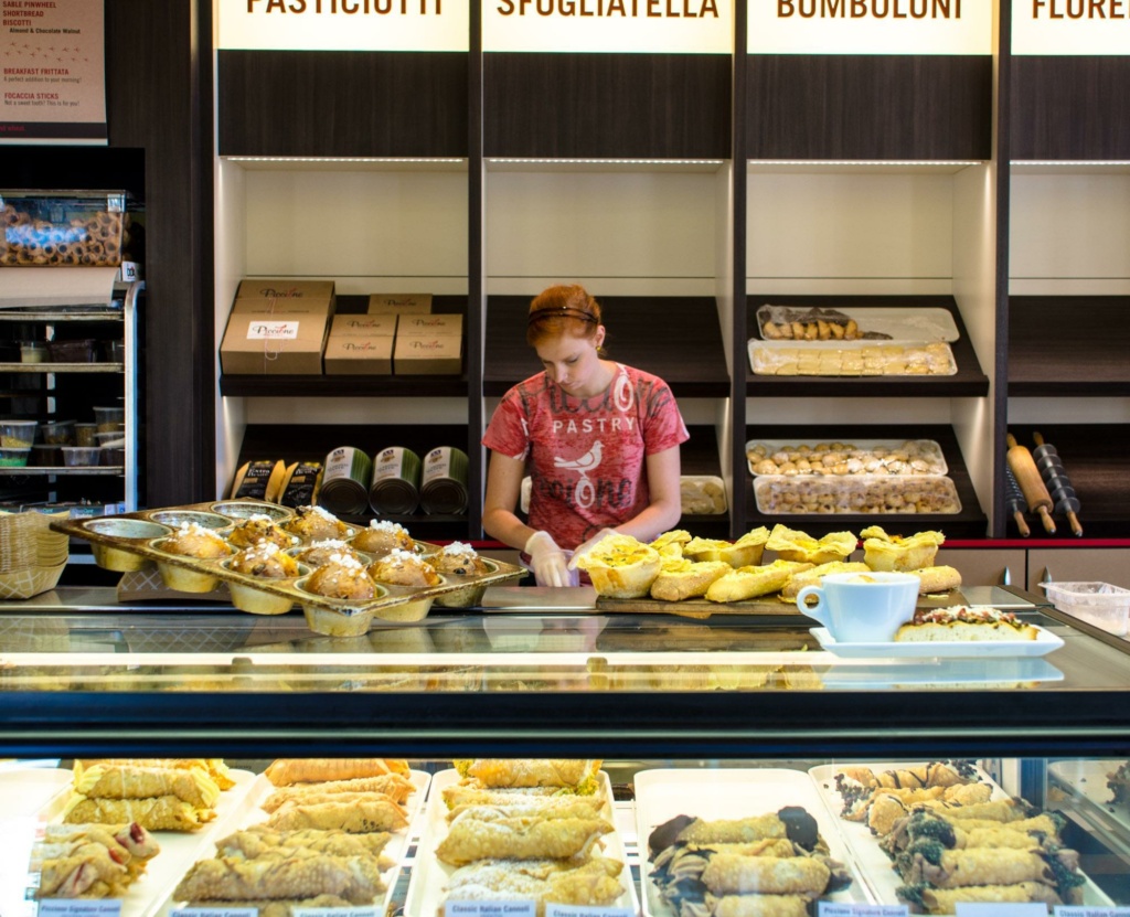 A person wearing a red shirt stands behind a counter filled with various pastries in a bakery. Shelves in the background display more baked goods, with a menu above listing items like "pasticciotti" and "bomboloni." A coffee cup sits on the counter.