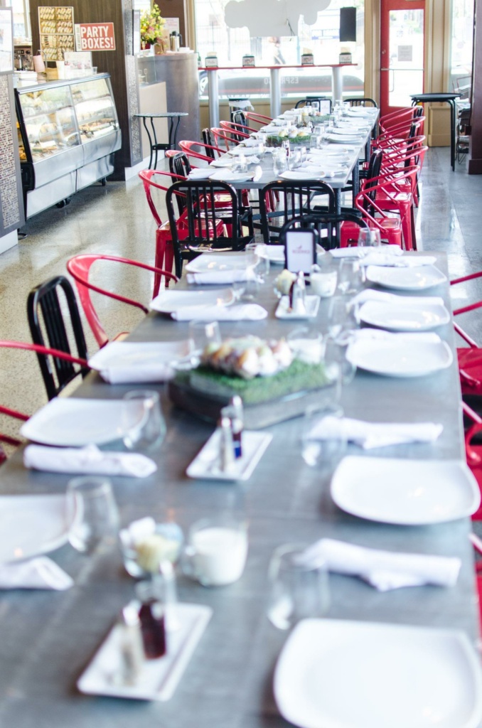 A long table set for dining with white plates, glasses, and napkins. Red and black chairs line both sides. In the background, a display case with desserts and a sign that reads "Party Boxes!" are visible. The ambiance is bright.