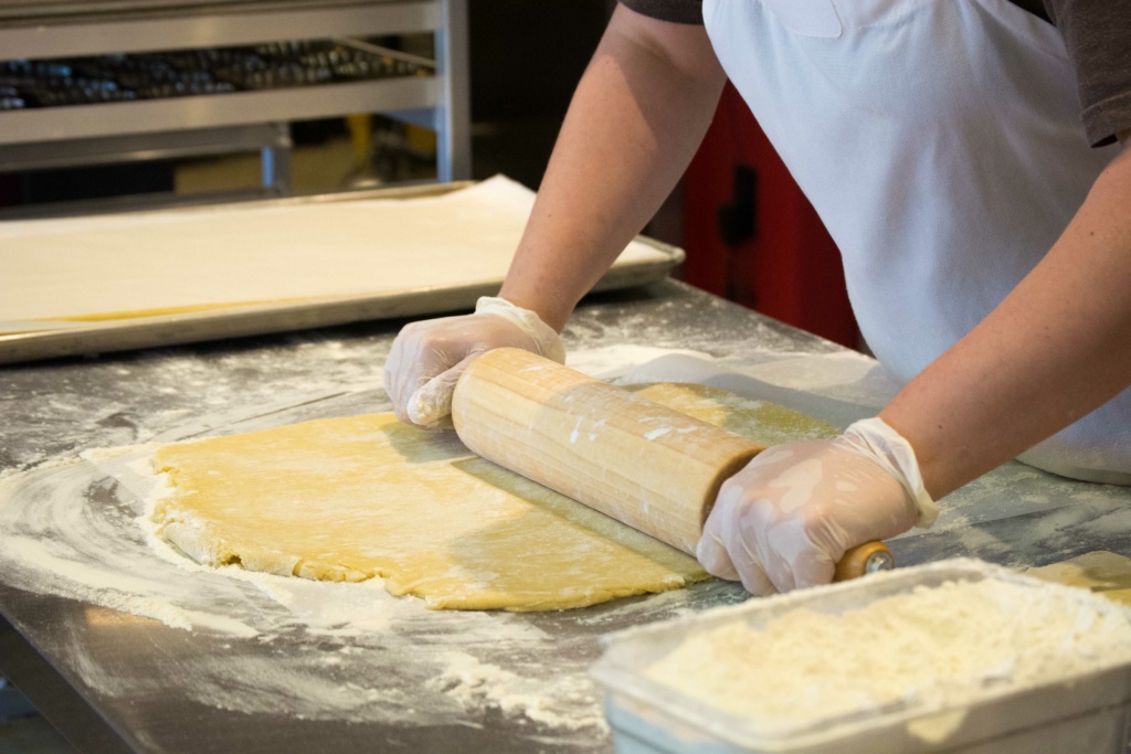 A person in an apron and gloves rolls out dough with a wooden rolling pin on a floured metal surface. A baking tray and container of flour are visible nearby.