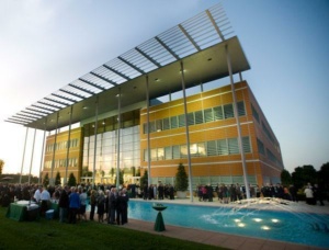 A modern building with large glass windows and a decorative metal rooftop is seen at sunset. In the foreground, a clear blue rectangular pool features water jets. A crowd of people gathers outside, creating a lively atmosphere.