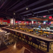 A spacious gaming facility with numerous poker tables and chairs arranged in rows. The room has dim lighting with red and purple accents. The ceiling features exposed beams, and several monitors are hung around the room.