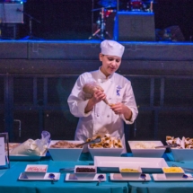 A chef dressed in a white uniform and hat is decorating desserts with a piping bag at a table. Various pastries and toppings are arranged around, and informational signs are placed on the table. Musical equipment is visible in the background.