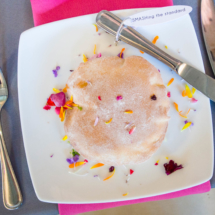 A round, powdered dessert on a plate, garnished with colorful flower petals. A fork and knife rest beside it, and a tag reading "SMASHing the standard" is tied to the knife. The table setting includes a pink napkin.