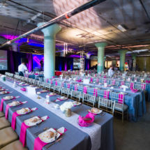 A large indoor event space features rows of elegantly set long tables with gray and pink tablecloths. Plates, glasses, and napkins are neatly arranged. A large screen displays "Kinesis" among other text, and ambient purple lighting creates a festive atmosphere.