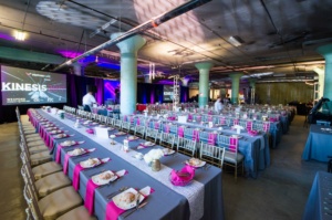 A large event space with multiple long tables set for a banquet. Tables are covered with gray tablecloths and decorated with pink runners, white plates, and floral centerpieces. A presentation screen in the background displays "Kinesis" and other logos.