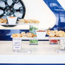 A snack table featuring crackers, pita bread, and breadsticks served with various spreads in white bowls. Decorative jars with vegetables are placed underneath, and a bottle of olive oil is on the right. The background has a blue and white design.
