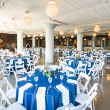 A spacious banquet hall is set for an event, with round tables covered in blue tablecloths and white napkins. Elegant glassware and floral centerpieces adorn the tables. Hanging paper lanterns provide a soft, warm light.