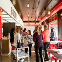 A group of women are gathered inside a brightly lit cafe, chatting and standing near a cart with drinks. The cafe has red accents, a high ceiling with hanging lights, and large windows letting in natural light.
