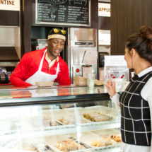 A man in a red jacket and apron serves pastries to a woman at a bakery counter. The woman is wearing a black and white checkered top. A menu board is visible in the background above the glass display case filled with pastries.