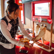 A woman is seen in a restaurant preparing a dessert by piping fillings into pastry cones. The setup includes labeled piping bags and dishes with various fillings like chocolate chips. Sunlight is streaming through the window, illuminating the scene.