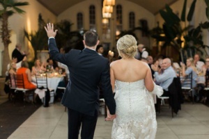 A bride and groom holding hands face a seated audience at a wedding reception. The groom waves with his other hand as the guests, seated at round tables, look on. The venue is elegantly decorated with plants and soft lighting.