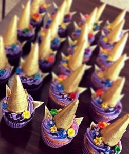 A group of cupcakes with purple frosting, topped with golden unicorn horn decorations, colorful flowers, and edible ears. They are arranged neatly on a dark surface.