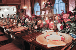 A lavishly decorated banquet table with elegant place settings and menus on gold chargers. The table is adorned with vibrant floral arrangements, candles, and soft lighting, creating a warm and inviting atmosphere in the luxurious venue.