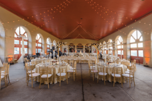 Spacious wedding reception hall with a red ceiling, adorned with string lights. Round tables are elegantly set with gold chairs and white tablecloths. Large windows line the walls, allowing natural light to fill the room.