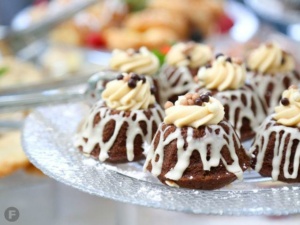 Mini bundt cakes topped with cream frosting and chocolate pearls are displayed on a glass platter. The cakes are drizzled with white icing, and there are blurred pastries in the background.