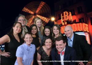 A group of people smiling in front of a backdrop featuring the iconic windmill of the Moulin Rouge, Paris. They are dressed in formal attire, with a mix of men and women posing together.
