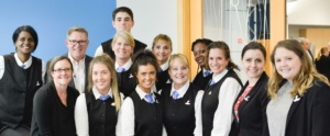 A group of twelve people smiling and posing together. They are wearing similar uniforms, including black vests and white shirts, with most having blue neckties. The background features a blue and wooden wall.