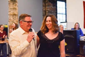 A man holding a microphone smiles while standing next to a woman at an indoor gathering. People are seated in the background, engaged and smiling. The setting has brick walls and large windows.