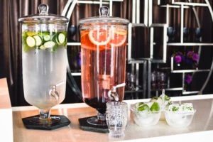 Two glass beverage dispensers on a counter; one with cucumber water, the other with grapefruit and berry water. Beside them are two glasses and two bowls of ice cubes with herbs and citrus slices.