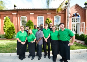 A group of restaurant staff members, wearing green shirts and black aprons, stand together smiling in front of a brick building with arched windows and palm trees in the background.