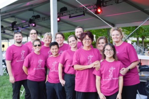 A group of people wearing matching pink shirts with a "Butler's Pantry" logo stand together and smile under a tent at an outdoor event. There are trees and tables in the background.