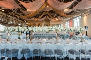 A beautifully decorated banquet hall features long tables with white linens, gold chargers, and elegant floral centerpieces with white flowers. Sheer fabric drapes are artfully hung from the ceiling, creating a sophisticated ambiance.