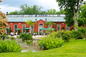 A brick greenhouse with large arched windows is surrounded by lush greenery and a small pond in the foreground. People sit at tables with green umbrellas on a sunny day.