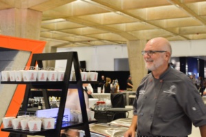 A smiling chef in a grey uniform stands beside a black display rack filled with white takeout boxes in a large venue. The setting has a modern, industrial feel with concrete pillars and bright lighting.